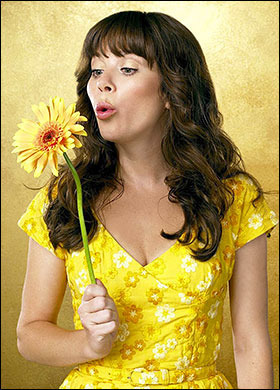 Anna Friel ve žlutých šatech fouká na květinu, kterou drží v ruce